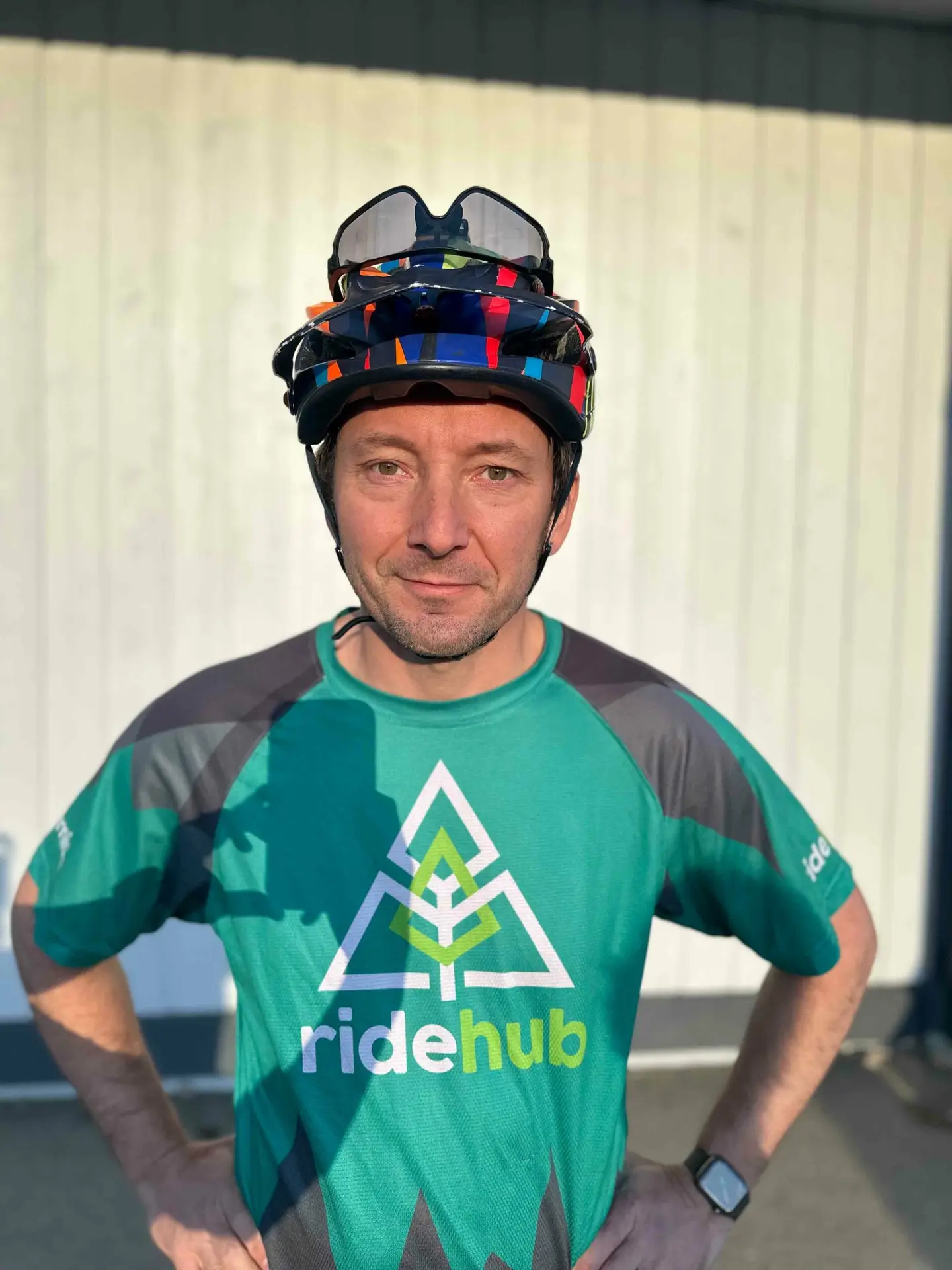 Meet the Team 11 Man in a bicycle helmet and green cycling jersey standing in front of a metal wall, the sun casting shadows on his face.