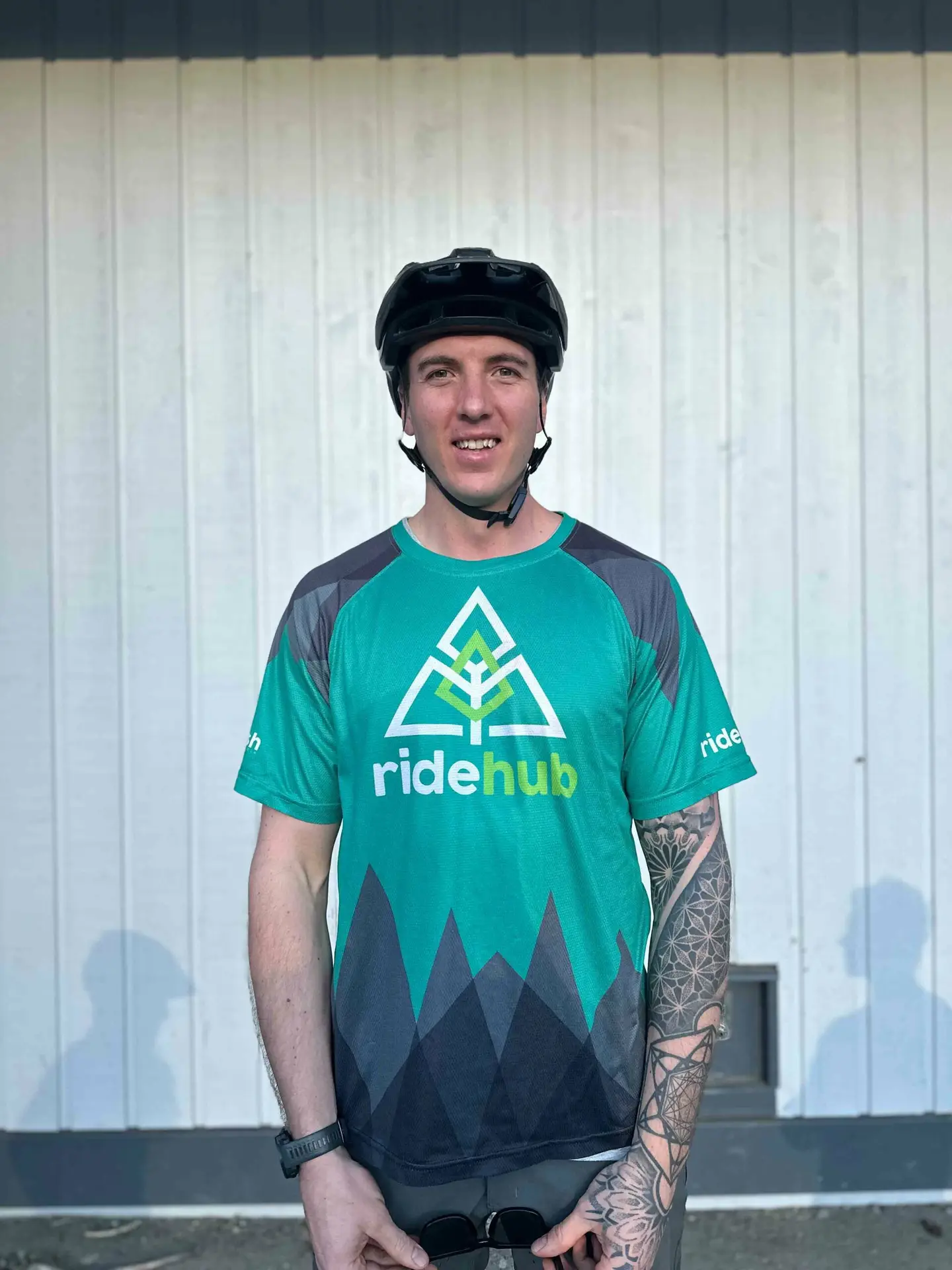 Meet the Team 9 Man in a green and blue ridehub jersey and black helmet standing in front of a white paneled wall, smiling at the camera.