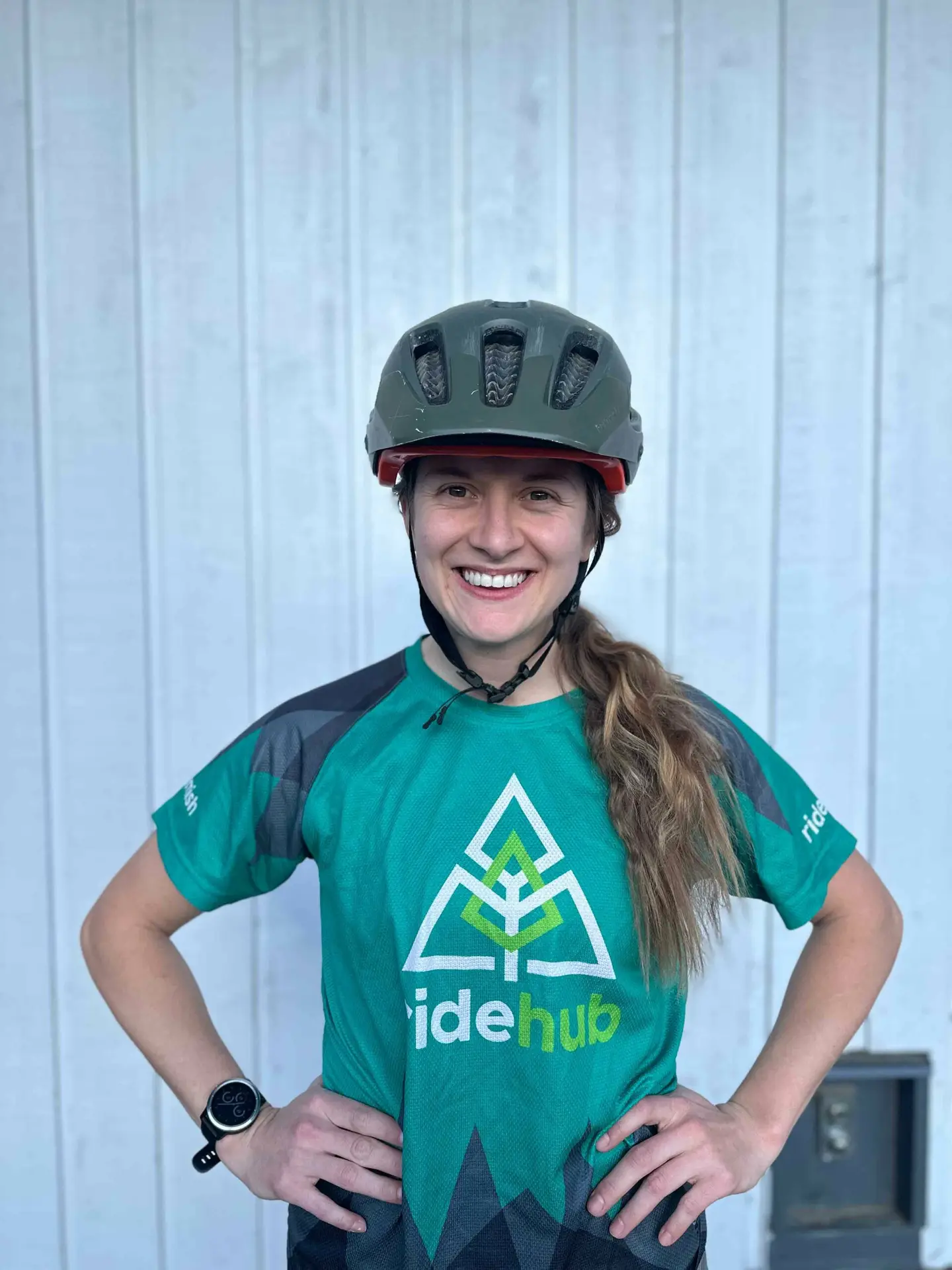 Meet the Team 7 Woman in a bike helmet and blue-green jersey smiling in front of a white corrugated metal background.