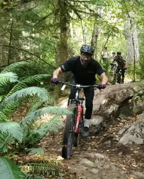 Meet the Team 8 A mountain biker navigates rocky terrain in a lush, green forest, followed by another rider, with a bystander watching from the side.