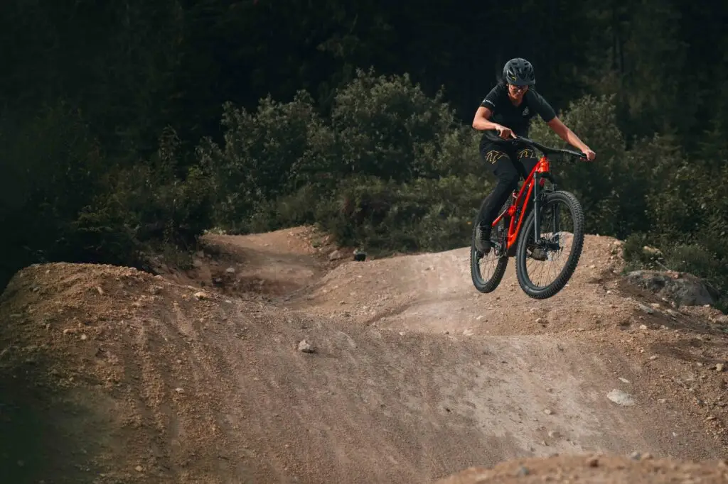A cyclist wearing a black shirt and helmet rides a red mountain bike, catching air on a dirt trail with a forested backdrop.