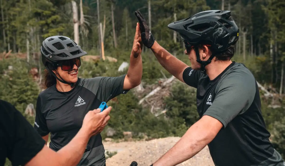 Private Instruction 2 Two cyclists in helmets and sport attire high-five each other in a wooded outdoor setting.