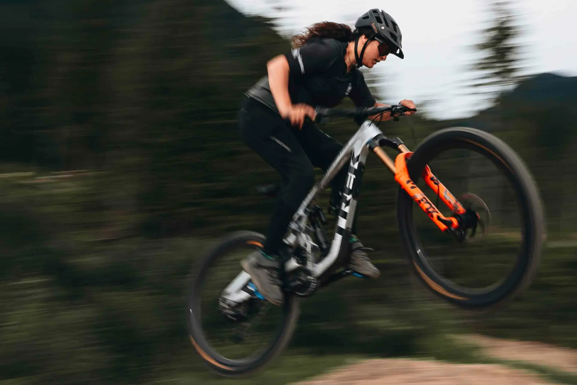 A person wearing black riding gear and a helmet is performing a jump on a mountain bike in a forested area.