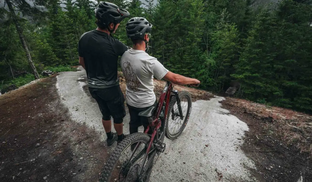 Private Instruction 3 Two cyclists wearing helmets stand on a winding trail in a forest, one with a red mountain bike. They appear to be observing the path ahead.