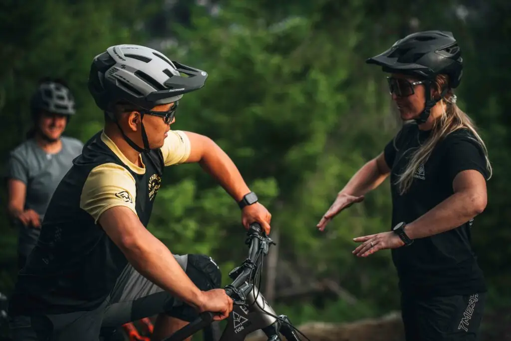 Shop 3 A person in cycling gear listens attentively to another person while holding their bike in a forested area with another cyclist in the background.