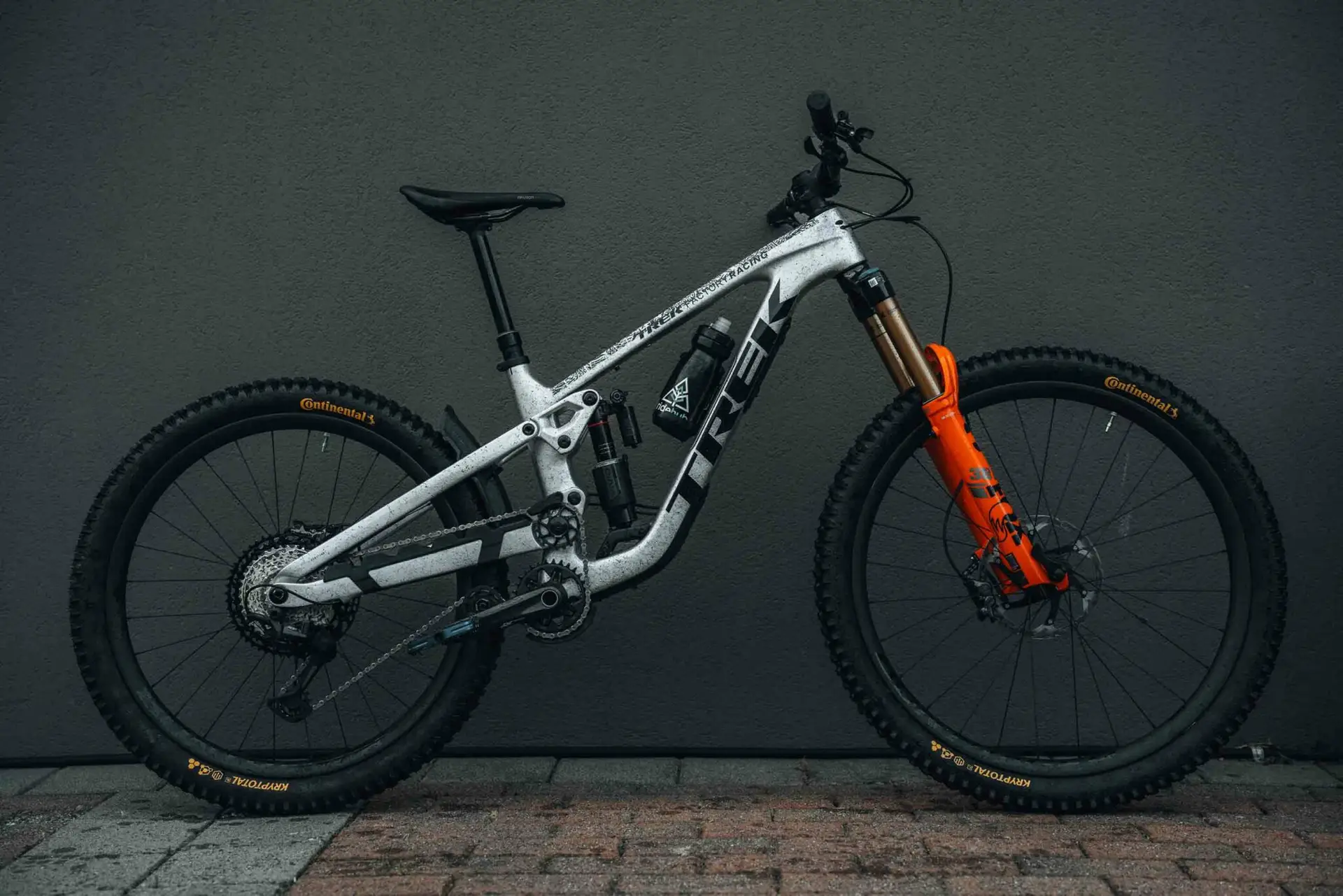 Shop 2 A gray Trek mountain bike with orange front suspension and black tires is positioned against a dark gray wall.
