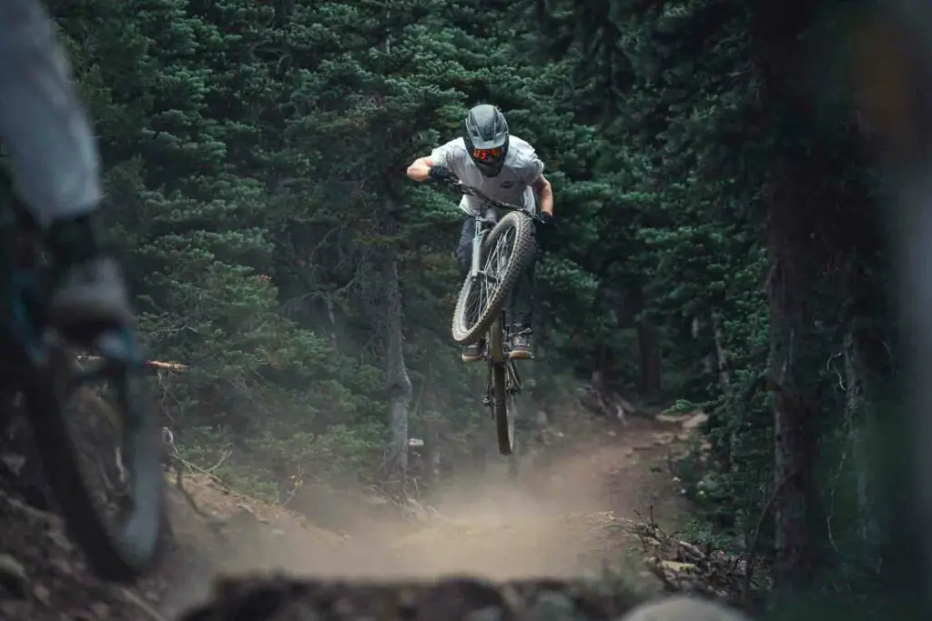 A mountain biker, wearing a helmet, jumps off a dirt trail in a dense forest, surrounded by trees and kicking up dust.