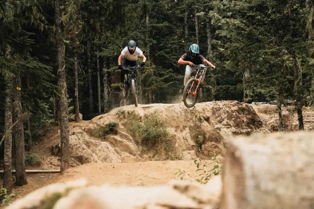 Two mountain bikers in helmets and gear are airborne, mid-jump, on a rocky downhill trail in a dense forest.