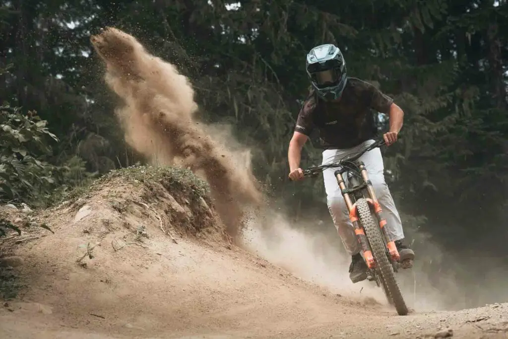 A mountain biker wearing a helmet rides down a dirt trail, kicking up dust behind them with trees in the background.