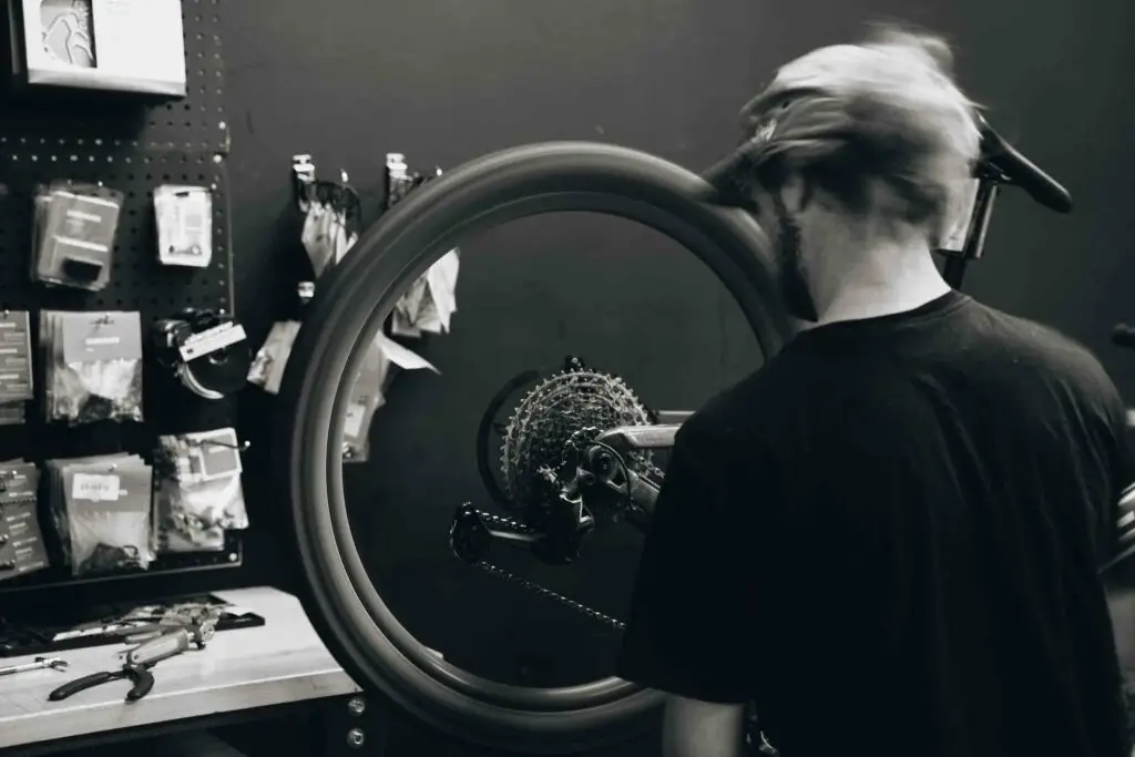 Shop 4 A person wearing a cap works on a bike wheel in a workshop, with various tools and parts hanging on a pegboard in the background.