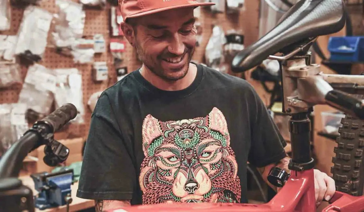 Shop 7 A person wearing a red cap and graphic t-shirt is assembling a red Trek bicycle in a workshop filled with bike parts and tools.