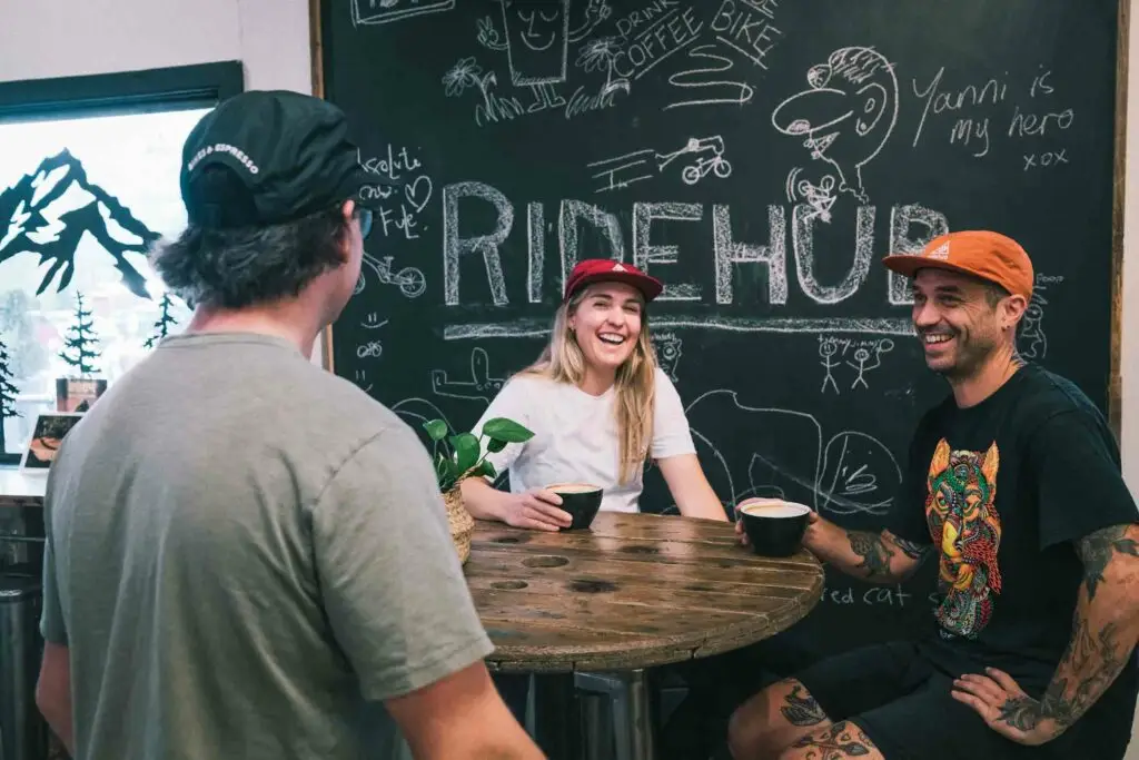 Shop 1 Three people sit at a round wooden table, drinking coffee and smiling in front of a chalkboard with doodles and