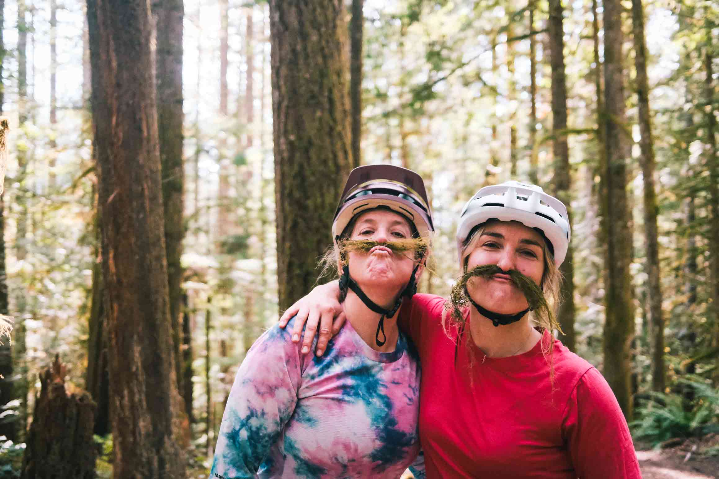 Shop 8 Two people wearing bike helmets and tie-dye shirts, posing playfully with moss under their noses in a lush forest near Squamish. It's a perfect moment capturing the fun spirit of mountain biking adventures in the shadow of breathtaking mountains.