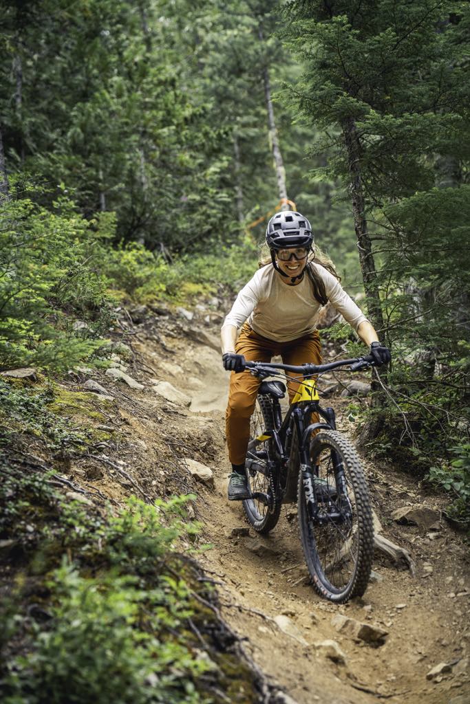 Meet the Team 10 A person wearing a helmet and gloves rides a mountain bike down a narrow, rocky forest trail surrounded by greenery.