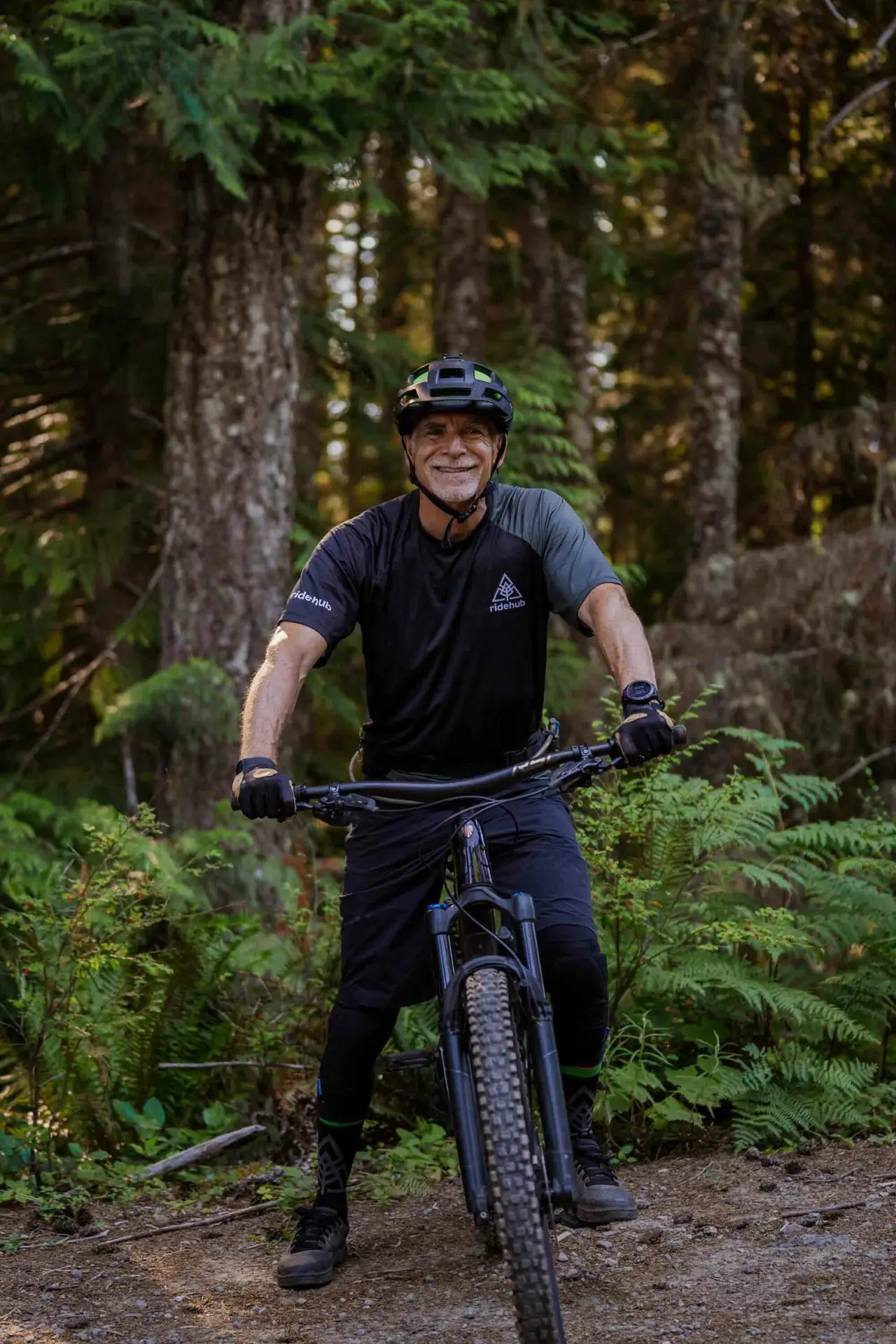 Meet the Team 6 A person wearing a helmet and cycling gear is smiling while sitting on a mountain bike in a forested area.