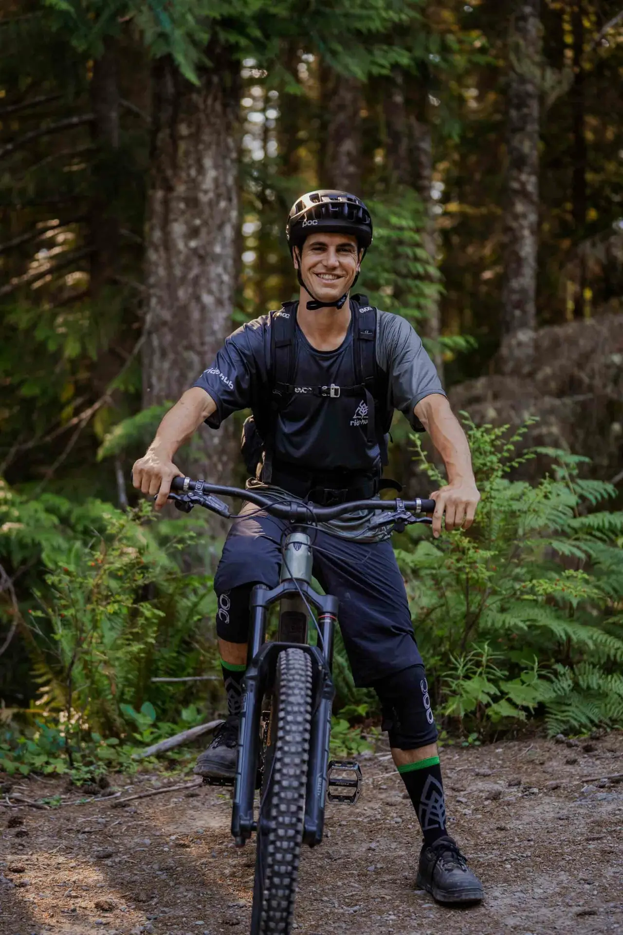 Meet the Team 4 A person wearing a helmet and sports gear is sitting on a mountain bike on a dirt trail in a forested area, smiling at the camera.