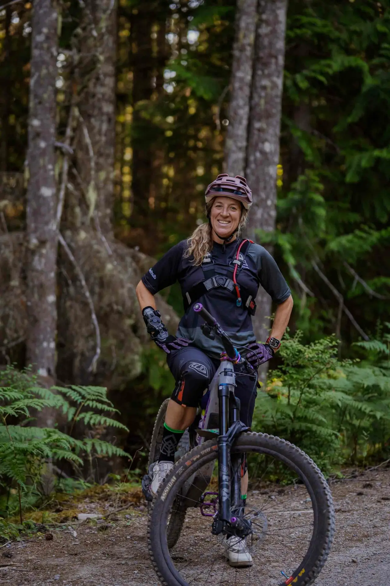 Meet the Team 1 A woman wearing protective gear stands with her mountain bike on a forest trail, smiling at the camera.