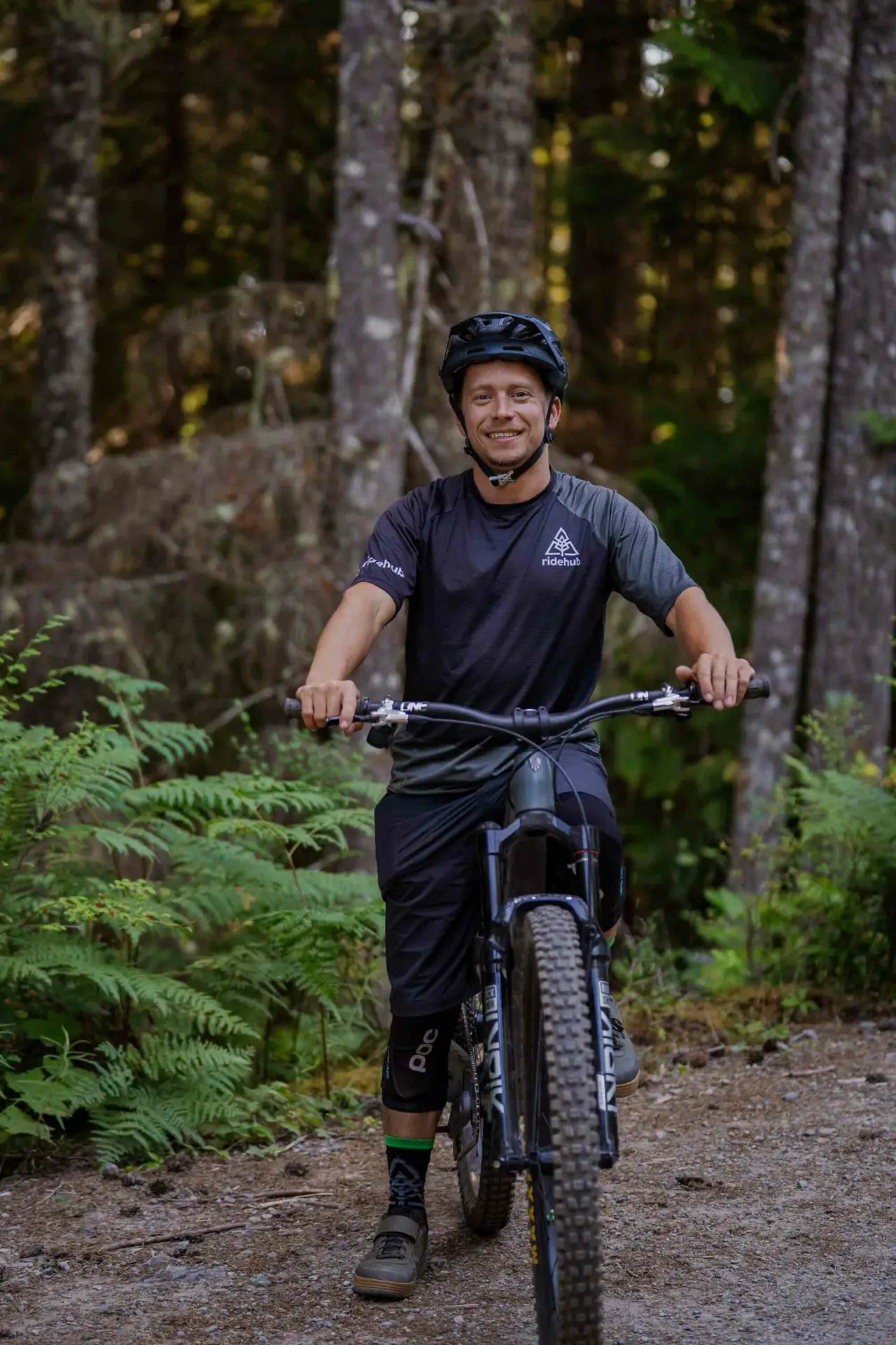 Meet the Team 3 A person wearing a helmet and cycling gear stands with a mountain bike on a forested trail.