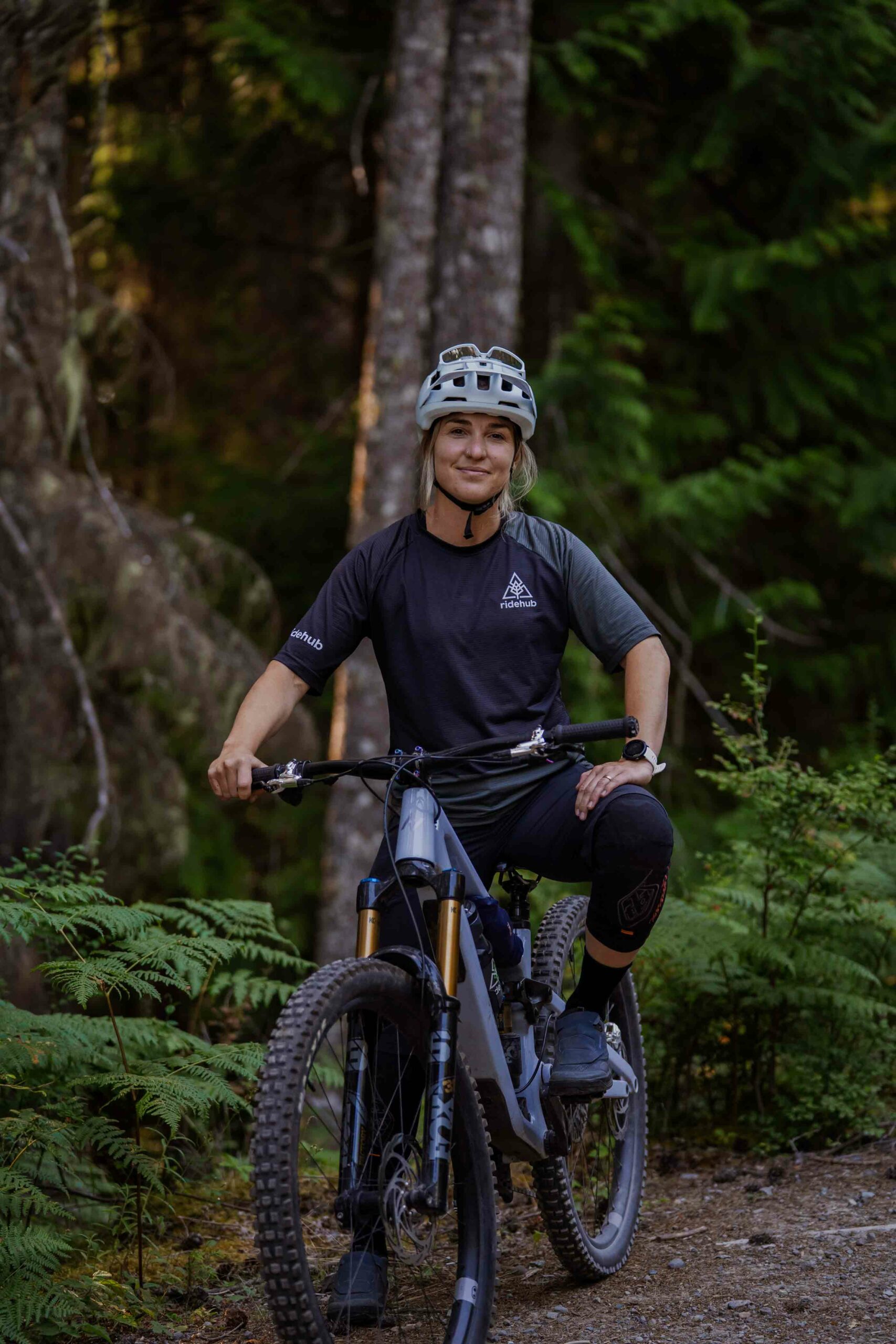 Meet the Team 2 Person wearing a helmet and biking gear sits on a mountain bike on a forest trail surrounded by trees and ferns.